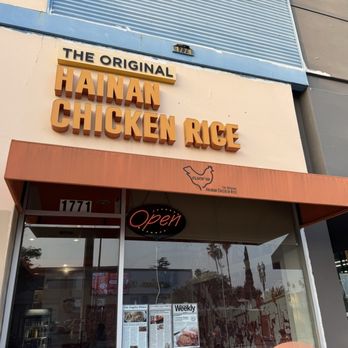 Cluck2Go - Exterior - The photo captures the exterior of a restaurant specializing in Hainan chicken rice, featuring a prominent sign with the text 'The Original Hainan Chicken Rice' and a smaller sign indicating 'Hainan' in bold letters. An illuminated 'Open' sign is visible below the main sign, and the storefront has a glass door with the street number 1771 displayed above. The building's facade includes a mix of beige and gray tones, with a blue awning or panel at the top. Reflections of palm trees and nearby structures can be seen in the glass window, indicating an outdoor urban setting. - Singaporean Near Me - Pasadena, California