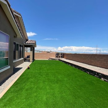 AMG Landscape - View of the yard, showing the block wall and planter areas. - A backyard features a vibrant green artificial lawn, a house with windows on the left, and a brick wall at the far end under a clear blue sky. - Irrigation Near Me