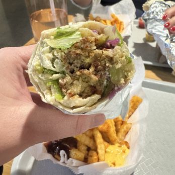 Kush - A hand holds a partially eaten burrito containing lettuce, meat, and other ingredients, with a tray of fries and dipping sauce visible on the table. In the background, other diners are seen, including a person using a smartphone. The setting appears to be a casual restaurant or fast-food establishment with a wooden table and food items served on trays. - Middle Eastern Near Me - Washington, DC, District of Columbia