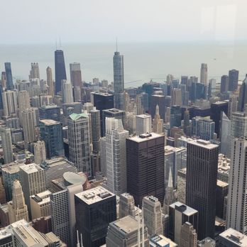 Skydeck Chicago - An aerial photograph captures the dense skyline of Chicago, Illinois, with a variety of tall buildings and skyscrapers. The image shows the urban landscape extending towards Lake Michigan, which is visible in the background. The photograph emphasizes the height and concentration of the city's architecture, with some buildings under construction and others with distinctive rooftop designs. - Observatories Near Me - Chicago, Illinois