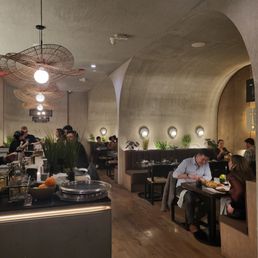 Main dining area - The image shows a restaurant interior with several diners seated at tables, engaged in conversations and dining. The decor includes modern, circular lighting fixtures hanging from the ceiling, a curved wall with small round lights, and potted plants along the wall. The space has a neutral color scheme with beige and brown tones, wooden flooring, and contemporary design features. The atmosphere appears relaxed and stylish, with a mix of booth seating and individual tables.