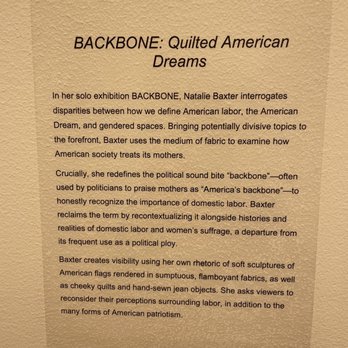 MOWA - St Kate - A beige textured background features a printed text panel providing an overview of Natalie Baxter's solo exhibition 'BACKBONE: Quilted American Dreams.' The text explains Baxter's interrogation of disparities in American labor, gendered spaces, and domestic labor through fabric art. It details her use of soft sculptures, quilts, and jean objects to challenge perceptions of patriotism and labor, emphasizing her focus on political and social themes. - Art Galleries Near Me - Milwaukee, Wisconsin