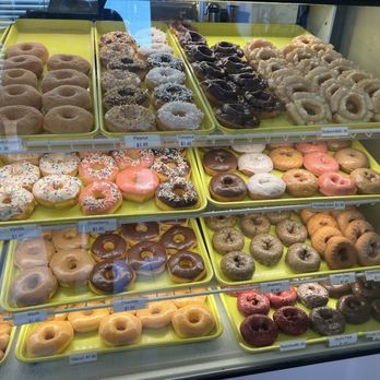 Texas Donuts - The image shows a glass display case containing multiple trays of donuts and baked goods. The donuts include glazed, chocolate, sprinkled, and specialty varieties such as blueberry, maple, and cinnamon sugar. Each tray is labeled with the flavor and price, and the donuts are arranged in neat rows. The display is part of a donut shop offering a diverse selection of freshly baked donuts and kolaches. - Donuts Near Me - Lorton, Virginia