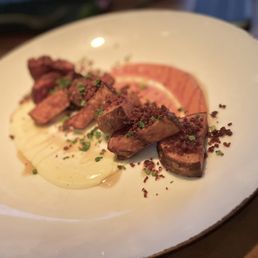 Patatas Bravas - A white ceramic plate holds slices of cooked meat, accompanied by a portion of mashed potatoes and garnished with chopped herbs. The presentation is simple, with the food arranged neatly on the plate, likely served at a restaurant setting.