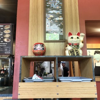 Toshi's Ramen - Japanese-style decorations, including a Maneki-neko and a Daruma doll, are placed on a wooden shelf in a restaurant interior, with a framed artwork above. - Ramen Near Me - Eugene, Oregon