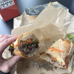 Bahn Mi - A hand holds a partially eaten sandwich filled with vegetables and meat, with another sandwich on paper in front, on a dark table.