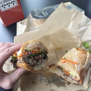 Le Croissant - Bahn Mi - A hand holds a partially eaten sandwich filled with vegetables and meat, with another sandwich on paper in front, on a dark table. - Bakeries Near Me - Logan, Utah