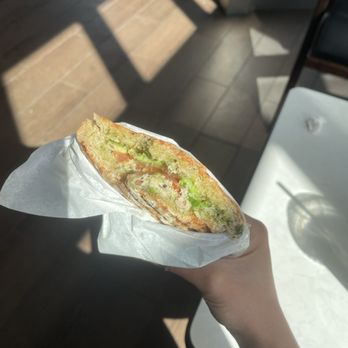 Revitasize - Tunacado sandwich - A person holds a sandwich wrapped in white paper, featuring avocado slices and tomato inside toasted bread. The background shows a tiled floor with sunlight and shadows, along with part of a white table and a chair, indicating an indoor setting with natural light. - Juice Bars & Smoothies Near Me - Markham, Ontario