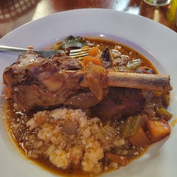 Vinny's Ristorante - Lamb osso bucco! Literally falls off the bone. - A white plate holds a serving of lamb osso bucco with a large bone, accompanied by rice and cooked vegetables in a broth. The dish appears to be a hearty, savory meal, likely served at an Italian or Sicilian-style restaurant. - Italian Near Me - Somerville, Massachusetts