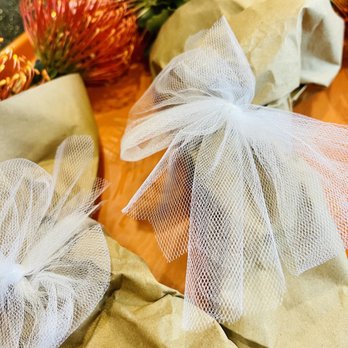 Daily Blooms - Beautiful free pin cushions with hand wrap - The image shows flower bouquets wrapped in brown paper, each tied with a white tulle bow. A pineapple with green leaves is visible in the background, placed on a dark, textured surface. The focus is on the wrapped flowers and bows, with the pineapple providing a colorful backdrop. - Home Decor Near Me - Fallbrook, California