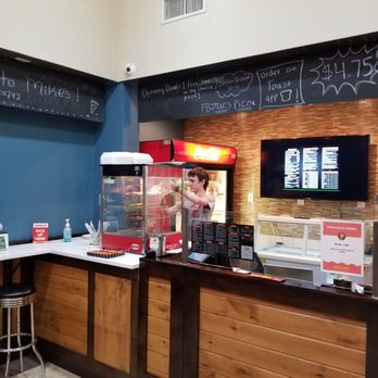 Mike's Pizza - a man ordering food at the counter - The image depicts the interior of a pizza restaurant counter area. A staff member is visible behind a glass display case, preparing food. The counter has a wooden paneling design, and there is a digital menu screen mounted on a brick wall behind the counter. Overhead, black chalkboard signs display handwritten messages and promotions. The area includes a small seating or waiting space with a stool and a sanitizer bottle on the counter. The setting appears clean and organized, with a focus on takeout and dine-in service. - Burgers Near Me - Chicago, Illinois