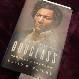 A hardcover biography of Frederick Douglass titled "Frederick Douglass: Prophet of Freedom" by David W. Blight, placed on a dark, plush fabric surface.