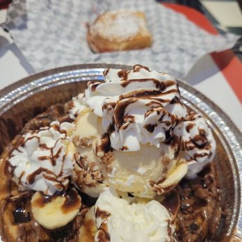 Chill...The Milkshake Bar - Nutella and banana waffle with Monte Cristo sandwich in background - A dessert bowl features a scoop of vanilla ice cream topped with whipped cream and chocolate syrup. Sliced bananas are placed around the ice cream, and additional chocolate syrup is drizzled over the whipped cream and bananas. The bowl is placed on a reflective metallic tray, with a waffle and a pastry in the background on a checkered paper, indicating a casual dining setting. - Ice Cream & Frozen Yogurt Near Me - Spring, Texas