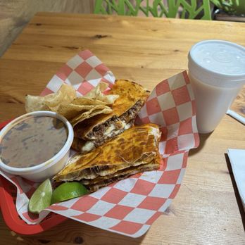 Taco Moro - Quesabirria Quesadilla - A quesabirria quesadilla served with a small bowl of beans, lime wedges, and a white drink on a wooden table at a restaurant. - Mexican Near Me - Chicago, Illinois