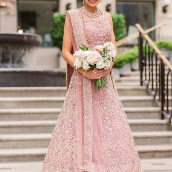 Makeup By Ana B - A woman wearing a pink embroidered traditional dress with intricate gold and silver details stands outdoors on steps, holding a bouquet of white and pink flowers. She is smiling and looking down, with a fountain and building with striped awnings in the background. She is accessorized with jewelry, including a necklace and earrings. - Makeup Artists Near Me