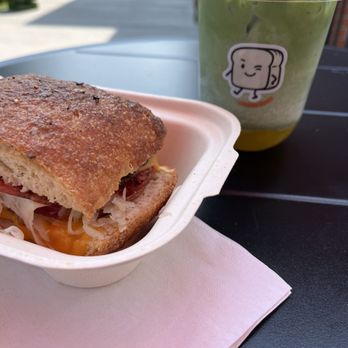 Moore Bread - Pastrami melt on ciabatta - A sandwich with bread, meat, and cheese in a takeout container, accompanied by a green drink with a cartoon sticker, on an outdoor table. - Bakeries Near Me - Denver, Colorado