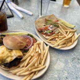 Burger in paradise and Philly sandwich - The image shows two plates of food on a table at a restaurant. Each plate contains a burger with cheese, French fries, and some vegetables such as pickles, onions, and tomatoes. There are also drinks, including a glass of iced soda with a straw and a glass of beer. The table surface is light-colored with a textured pattern, and there are napkins and utensils visible in the background.