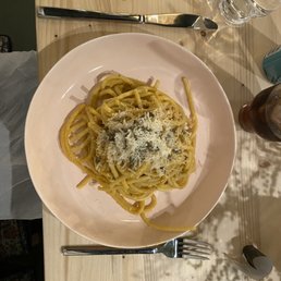 a plate of pasta with parmesan cheese - A white round plate contains a serving of spaghetti pasta topped with grated cheese, placed on a light-colored wooden table. The table also has a metal fork positioned below the plate, a glass of water, and a dark-colored drink in a glass bottle. The setting appears to be a casual dining environment.
