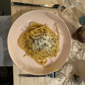 Mila's Mesa - a plate of pasta with parmesan cheese - A white round plate contains a serving of spaghetti pasta topped with grated cheese, placed on a light-colored wooden table. The table also has a metal fork positioned below the plate, a glass of water, and a dark-colored drink in a glass bottle. The setting appears to be a casual dining environment. - Caterers Near Me - Albuquerque, New Mexico