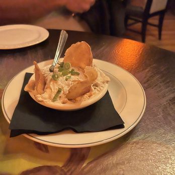 Sole's - Whiskey onion dip - A small white bowl contains potato salad topped with chopped green onions and two potato chips. The bowl is placed on a black napkin on a dark wooden table in a restaurant setting. In the background, a person is partially visible, and a glass of beverage is on the right side of the image. - Bars Near Me - Keene, New Hampshire