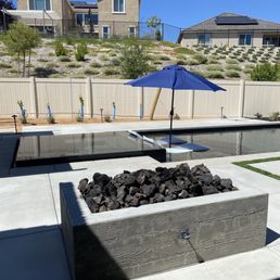 A backyard pool area features a black-tiled pool, a blue umbrella with lounge chairs, and a fire pit with rocks, enclosed by a white fence.