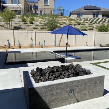 Bella Vista Pools - A backyard pool area features a black-tiled pool, a blue umbrella with lounge chairs, and a fire pit with rocks, enclosed by a white fence. - Pool & Hot Tub Service Near Me
