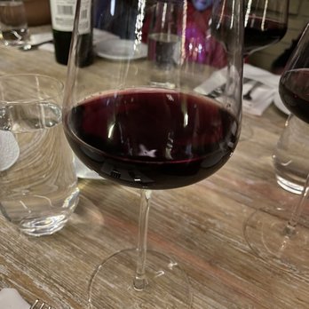 Cafe Central - Wine - A glass of red wine is placed on a wooden table, with other glasses, utensils, and a bottle visible in the background. - Salad Near Me - El Paso, Texas