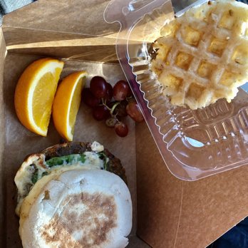 Velo Cafe - Egg muffin sandwich with pepper jack, spinach, walnut Patty, with fruit on the side and a specially made for me potato cake!!! - A breakfast meal includes an egg muffin sandwich with greens, orange slices, grapes, and a waffle in a plastic container, on a brown paper tray. - Juice Bars & Smoothies Near Me - Cable, Wisconsin