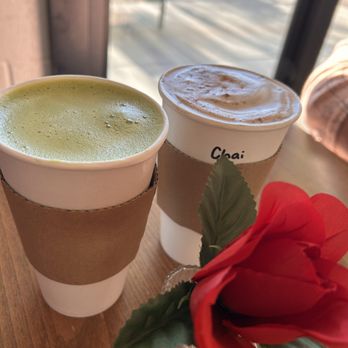 Collective Coffee - Matcha & chai lattes - Two coffee beverages, one with green matcha and the other with foam, sit on a wooden table near a window, with a red flower in a glass vase. - Coffee & Tea Near Me - Phoenix, Arizona