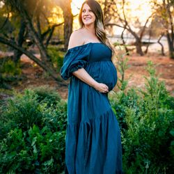 Madison Grey Photography - Pregnant woman wearing a blue off-the-shoulder dress standing outdoors at sunset, surrounded by trees and greenery.
