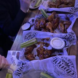 Large tray of celery, followed by chipotle bbq, honey bbq and spicy ranch (a dry rub) - Several baskets of chicken wings with celery sticks and dipping sauces are arranged on a wooden table, with a person partially visible eating in the background.