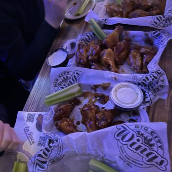 Big City Wings - Large tray of celery, followed by chipotle bbq, honey bbq and spicy ranch (a dry rub) - Several baskets of chicken wings with celery sticks and dipping sauces are arranged on a wooden table, with a person partially visible eating in the background. - Chicken Wings Near Me - Spring, Texas