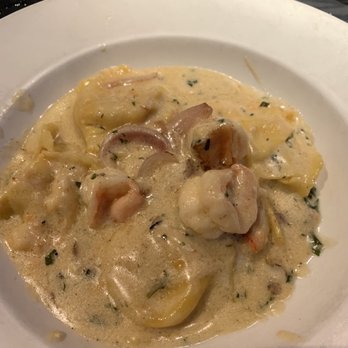 Bella Milano - Seafood Ravioli - The image shows a white, round plate with seafood ravioli covered in a creamy, herb-speckled sauce. The ravioli are filled with seafood, and pieces of shrimp and possibly other shellfish are visible. The dish is served on a dark table, with a glass of water and a basket in the background, indicating a restaurant setting. - Italian Near Me - Springfield, Illinois