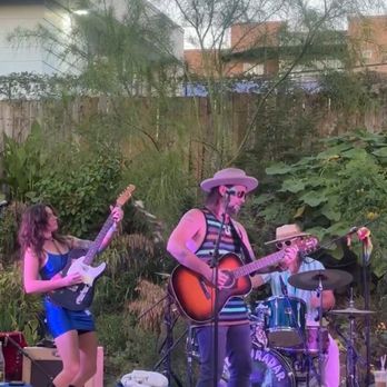 Cosmic - Live band - A live band performs outdoors on a stage set against a backdrop of trees and residential buildings. The band includes a female guitarist in a blue dress, a male guitarist and singer wearing a hat and striped shirt, and a drummer behind a drum kit. The stage has small tables with blue tablecloths, and audio equipment is visible in the foreground. The scene suggests a casual, outdoor music event. - Coffee & Tea Near Me - Austin, Texas