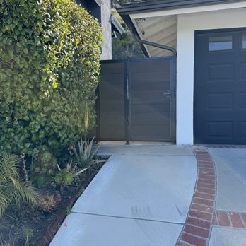 Socal Gates - A black gate is positioned beside a white building with a garage door, next to a green hedge and a concrete driveway with brick accents. - Fences & Gates Near Me - Orange, California