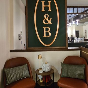 Haley & Bear - The image shows a cozy seating area with two orange upholstered chairs, each with a striped pillow, flanking a small round table. The table holds a decorative white vase, a small gold lamp, and other small objects. Behind the chairs, a large sign with the letters 'H & B' inside an oval frame is mounted on the wall. The background reveals part of a restaurant or cafe interior with modern lighting fixtures and a visible bar or counter area. - Breakfast & Brunch Near Me - Concord, New Hampshire