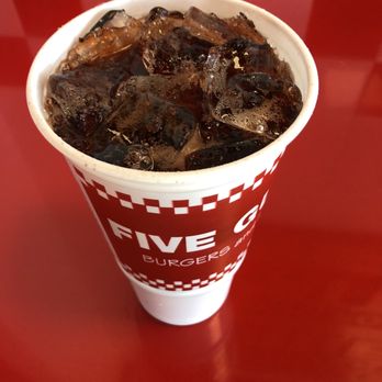 Five Guys - Cherry Dr Pepper - A disposable paper cup containing Cherry Dr Pepper soda with ice cubes is positioned on a red table. The cup has a red and white checkered pattern and the text 'FIVE GUYS BURGERS' visible on the side. The background features a red and white tiled wall, indicating the setting is inside a Five Guys restaurant in Santa Fe, New Mexico. - Burgers Near Me - Santa Fe, New Mexico