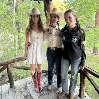 Rainbow Trout Ranch - Ready for Western Dancing - Three women wearing cowboy hats and western-style clothing stand on a wooden balcony surrounded by green trees in a forest. - Resorts Near Me - Antonito, Colorado