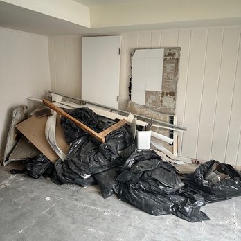 Fila Construction - Pile of rubble from bathroom demo that Lucas didn't remove from our home - Construction debris, such as black trash bags, wooden planks, metal pipes, and cardboard, is piled against a wall in an indoor space with white paneled walls and a concrete floor. The wall has a partially removed mirror or panel, revealing exposed wall material. The scene suggests ongoing renovation or demolition work. - General Contractors Near Me