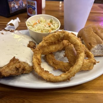 Oscar Store - Chicken Fried Steak, Onion Rings and Coleslaw - A restaurant meal featuring fried chicken with white gravy, served with onion rings, a small bowl of coleslaw, a bread roll, and a large drink in a to-go cup. The food is arranged on a white oval plate on a wooden table, with condiments and utensils visible in the background. - American Near Me - Temple, Texas