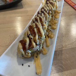 Crystal Roll - A sushi roll with several pieces arranged in a line on a long white plate. The roll is topped with mayonnaise, eel sauce, and black sesame seeds, with additional sauces drizzled over the plate. The background shows a wooden table and a blurred dish with sashimi or similar food. The setting appears to be a restaurant specializing in sushi and Japanese cuisine.