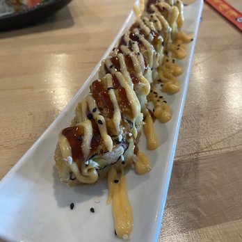 Kasabi - Crystal roll. (One of their most popular) - A sushi roll with several pieces arranged in a line on a long white plate. The roll is topped with mayonnaise, eel sauce, and black sesame seeds, with additional sauces drizzled over the plate. The background shows a wooden table and a blurred dish with sashimi or similar food. The setting appears to be a restaurant specializing in sushi and Japanese cuisine. - Ramen Near Me - Festus, Missouri