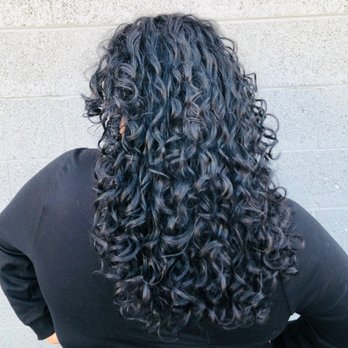 Salon Estique - A person with long, dark, curly hair is shown from the back, standing against a gray brick wall. They are wearing a black top. - Hair Salons Near Me - Phoenix, Arizona
