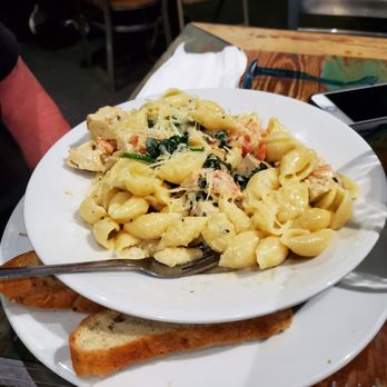 Cafe Eleven - Basil Chicken Pasta - A white plate contains basil chicken pasta with a creamy sauce, garnished with herbs and cheese, placed on a wooden table. Accompanying the pasta is a slice of garlic bread on a separate plate. The background shows chairs and part of the restaurant interior, indicating a dining setting. - Bubble Tea Near Me - Saint Augustine, Florida