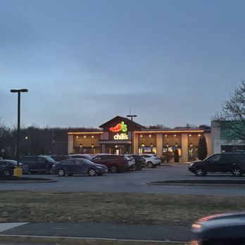 Chili's - The image shows the exterior of a Chili's restaurant with its illuminated sign featuring a chili pepper logo and the name 'Chili's'. The building is situated in a parking lot with multiple cars parked in front. The sky is transitioning from day to night, indicating dusk or early evening. The scene includes parking lot lighting and some surrounding trees and structures. - Bars Near Me - Raynham, Massachusetts