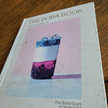 YumCha - The image shows the cover of 'The Boba Book' with the subtitle 'Bubble Tea and Beyond.' The cover displays a photograph of a layered bubble tea drink with three distinct layers: blue, white, and purple, with tapioca pearls at the bottom. The background of the cover is pink, and the book appears to be placed on a wooden surface. The book is authored by 'The Boba Guys' with Richard Parks III. - Bubble Tea Near Me - Waltham, Massachusetts