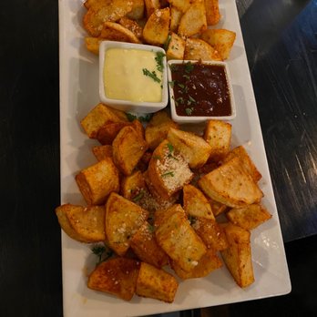 Debajo Tapas ABQ - Patatas Bravas - A rectangular white plate contains roasted potato chunks, seasoned and garnished with herbs. Two small bowls of dipping sauces, one yellow and one dark, are placed in the center of the plate. The dish is served on a dark table, with a glass of water visible in the background. - Spanish Near Me - Albuquerque, New Mexico