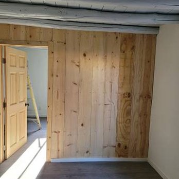 Always Construction Services - A room features a wooden wall made of vertical planks, with an open door on the left side leading to another space. The adjacent room has a wooden door and a ladder visible inside. The ceiling has exposed pipes or ducts, and the floor is covered with grayish flooring. The overall setting appears to be a construction or renovation site. - Stucco Services Near Me - Santa Fe, New Mexico