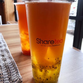 Sharetea - Peach kiwi green tea with aiyu jelly - Two large transparent cups of peach kiwi green tea with aiyu jelly are on a wooden table near a window. The drinks contain visible black seeds and pieces of fruit, with the Sharetea logo printed on the cups. The background shows a window with a view outside, and a cloth napkin is partially visible on the left side. - Bubble Tea Near Me - St. Charles, Illinois