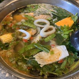 Bowl of  Shabu - A metal hot pot contains a broth with vegetables such as leafy greens, corn, and sliced squash, along with seafood including squid rings. The broth appears to be simmering, with bubbles and herbs floating on the surface. In the background, a small bowl of rice and a basket of greens are partially visible. The setting suggests a dining experience focused on hot pot cuisine.
